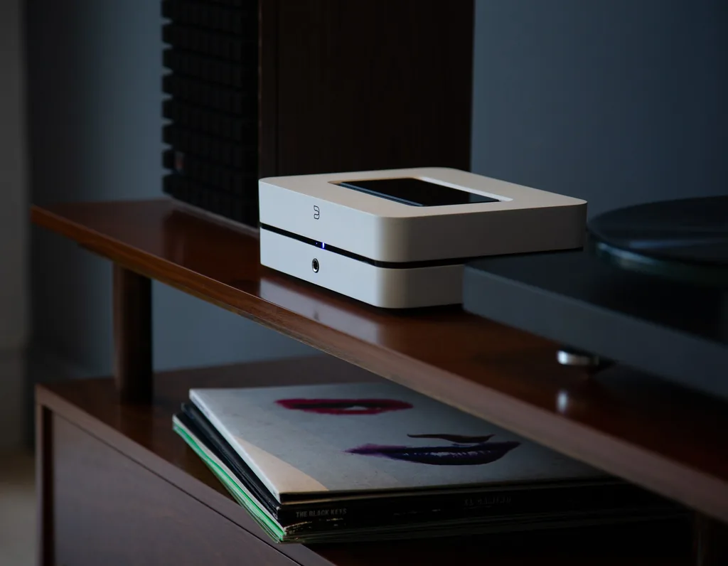 Amplificador com streaming Bluesound POWERNODE branco sobre móvel de madeira escura, ao lado de um toca-discos, com discos na prateleira de baixo