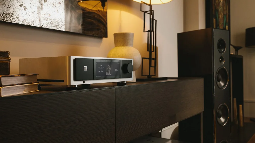 Amplificador integrado com streaming NAD Masters M33 V2 sobre aparador de madeira escura, ao lado de caixa acústica de torre, em sala com luz quente