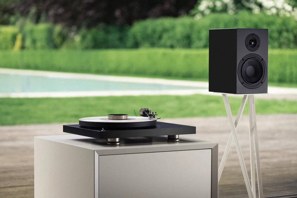 Toca-discos Pro-Ject Debut PRO sobre móvel branco, com caixa acústica em pedestal, numa varanda diante do jardim e da piscina