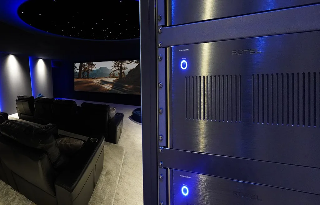Amplificadores multicanal Rotel da série RMB MKII instalados em rack, ao lado de uma sala de home cinema