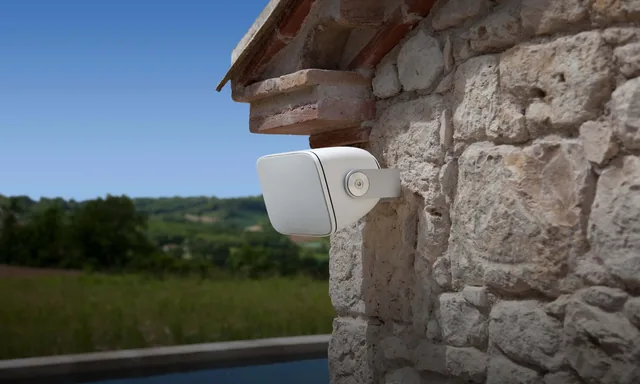 Caixa externa Bowers & Wilkins fixada em parede de pedra, com piscina e campo ao fundo